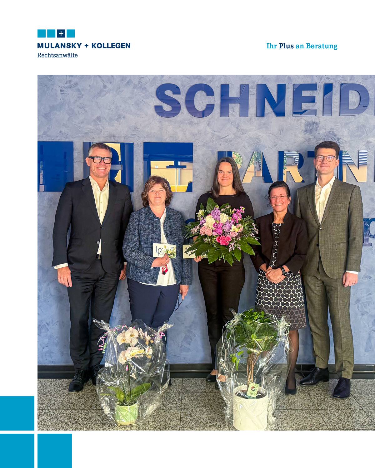Drei Frauen stehen zwischen zwei Herren, eine hält einen Blumenstrauß, zu Füßen der anderen weitere Blumenpräsente.